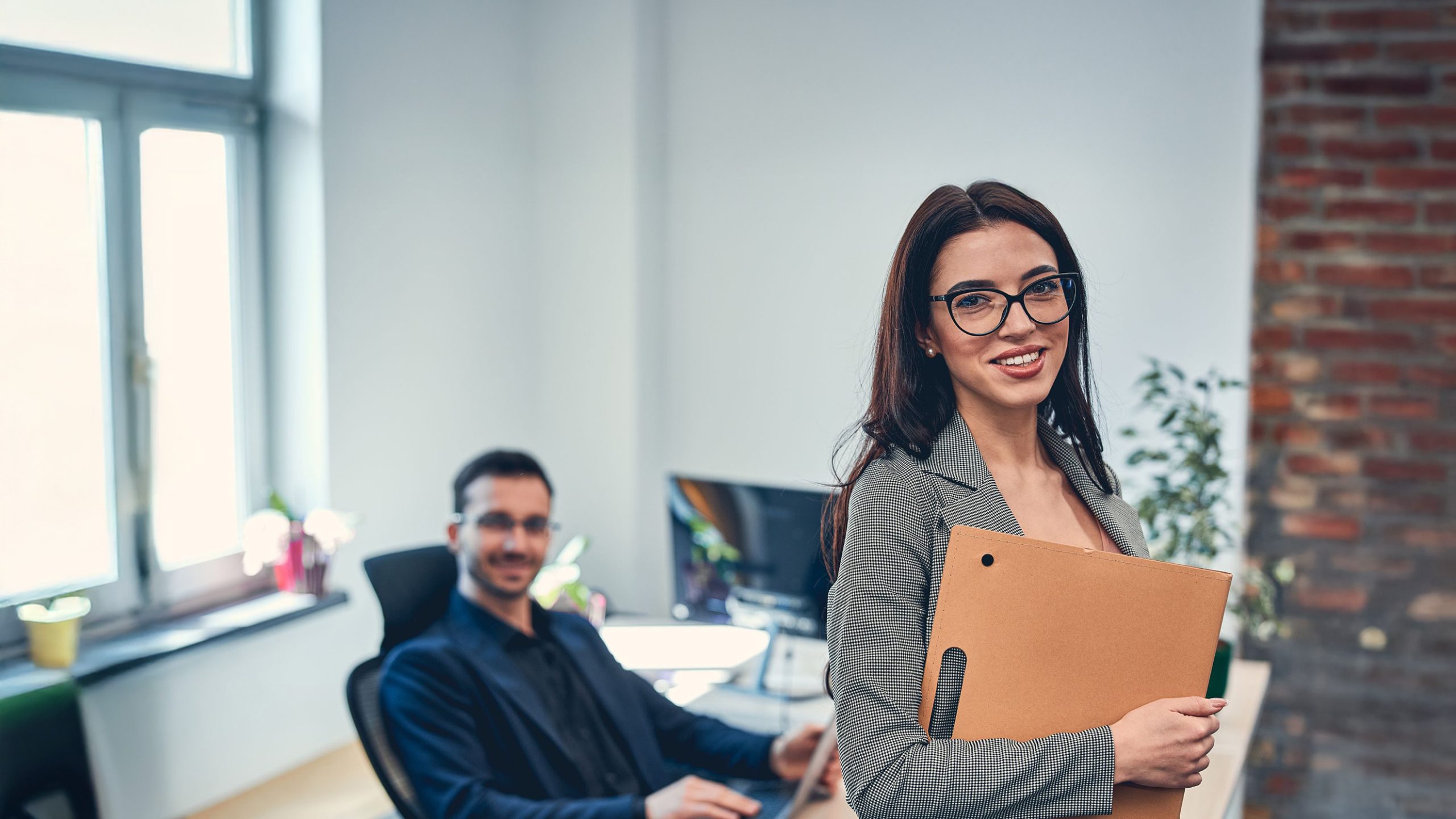 Backoffice medewerker met bril, lachend naar de camera; op de achtergrond zit een mannelijke collega.